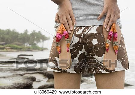 Stock Photo - Mid section view of a woman holding a pair of flip-flops on her back. Fotosearch