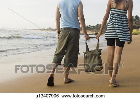 Rear view of a young couple holding a bag and walking on the beach View Large Photo Image Stock Photograph - Rear view of a young couple holding a bag and walking on the beach. Fotosearch
