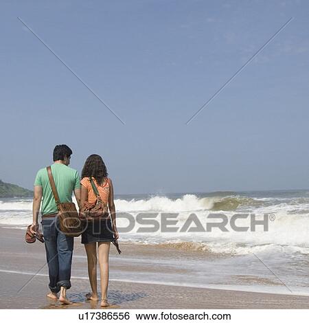 Stock Photograph - Rear view of a young couple holding their slippers and walking on the beach. Fotosearch