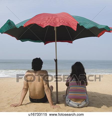 Stock Photography - Rear view of a young couple sitting under a beach umbrella on the beach. Fotosearch