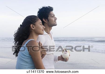 Stock Image - Young woman standing with a young man holding a glass of cocktail on the beach. Fotosearch