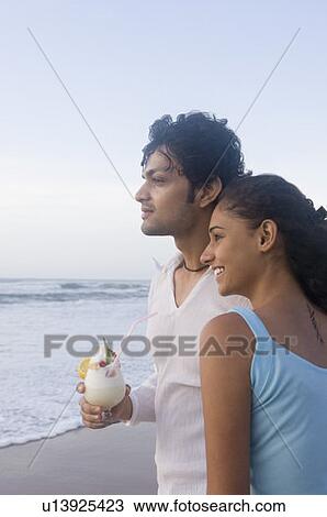 Stock Image - Young woman standing with a young man holding a glass of cocktail on the beach. Fotosearch