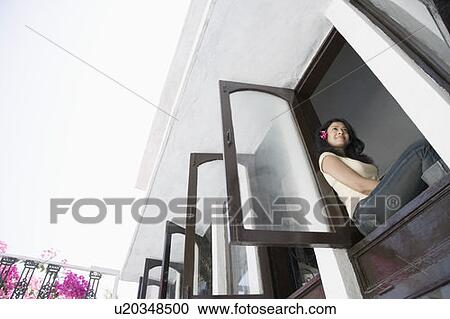 Stock Image - Low angle view of a young woman sitting at the ledge of a window. Fotosearch