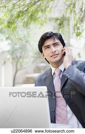 Businessman standing in front of a laptop and talking on a mobile phone View Large Photo Image Stock Photograph - Businessman standing in front of a laptop and talking on a mobile phone. Fotosearch