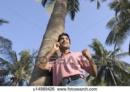 Stock Photograph - Low angle view of a young man leaning against a tree and talking on a mobile phone. Fotosearch