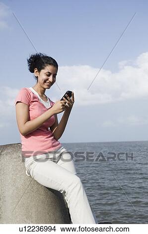 Young woman sitting on a rock and text messaging on a mobile phone View Large Photo Image Picture - Young woman sitting on a rock and text messaging on a mobile phone . Fotosearch