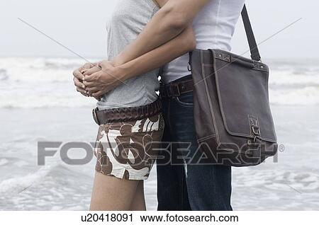 Stock Image - Mid section view of a man embracing a woman from behind on the beach. Fotosearch