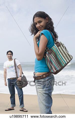 Stock Image - Portrait of a young woman carrying a hand bag with a young man standing in the background on the beach. Fotosearch