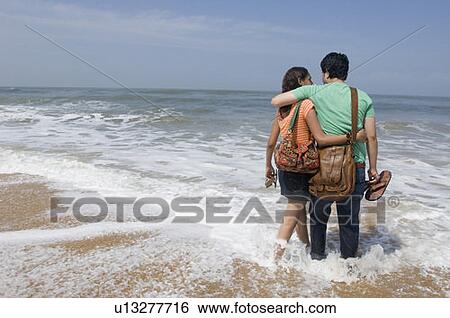 Stock Photograph - Rear view of a young couple walking on the beach with arm around each other. Fotosearch