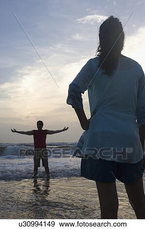 Rear view of a young woman standing on the beach with a young man standing in front of her with his arm outstretched View Large Photo Image Stock Photo - Rear view of a young woman standing on the beach with a young man standing in front of her with his arm outstretched. Fotosearch