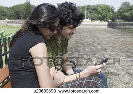 Side profile of a young couple sitting on a bench and operating a mobile phone, Goa, India View Large Photo Image Stock Image - Side profile of a young couple sitting on a bench and operating a mobile phone, Goa, India. Fotosearch