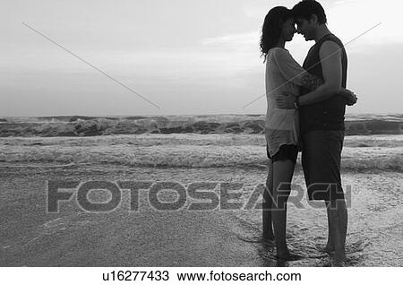 Stock Image - Side profile of a young couple standing on the beach and embracing each other. Fotosearch