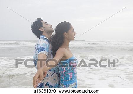 Stock Image - Side profile of a young couple standing with their arms outstretched on the beach. Fotosearch