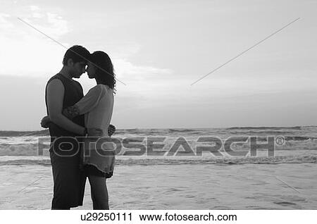 Stock Image - Side profile of a young couple embracing each other on the beach. Fotosearch