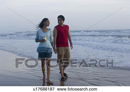 Stock Photo - Young man walking on the beach with a young woman holding a glass of drink. Fotosearch