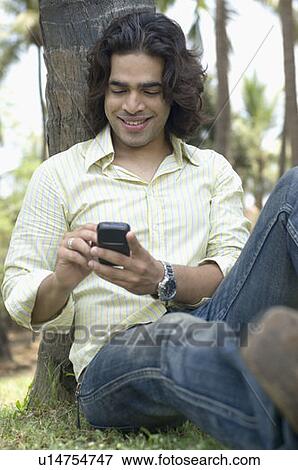 Close-up of a young man leaning against a tree trunk and text messaging with a mobile phone View Large Photo Image Stock Photo - Close-up of a young man leaning against a tree trunk and text messaging with a mobile phone. Fotosearch