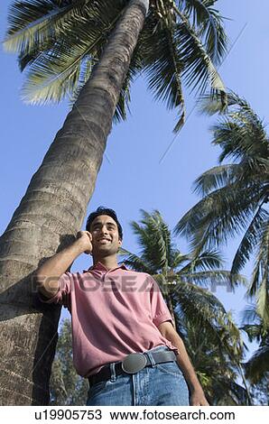 Stock Image - Low angle view of a young man leaning against a tree and talking on a mobile phone. Fotosearch