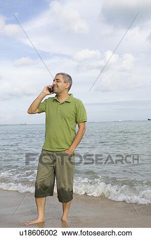 Stock Image - Mid adult man standing on the beach and talking on a mobile phone. Fotosearch