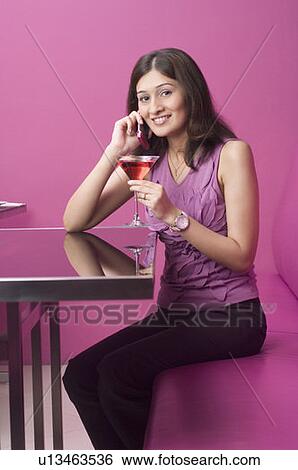 Stock Photograph - Portrait of a young woman talking on a mobile phone with holding a martini glass. Fotosearch