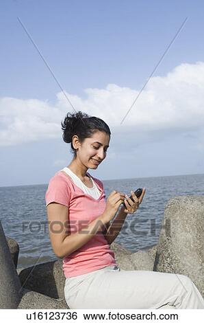 Stock Photograph - Side profile of a young woman sitting on a rock and text messaging on a mobile phone . Fotosearch