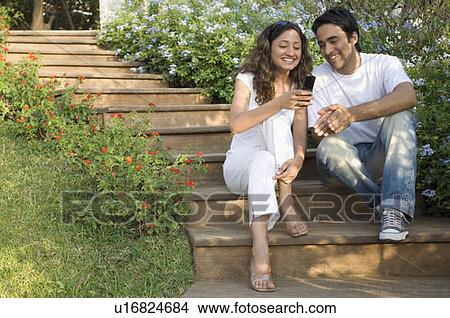 Young couple sitting in a garden and looking at a mobile phone View Large Photo Image Picture - Young couple sitting in a garden and looking at a mobile phone. Fotosearch