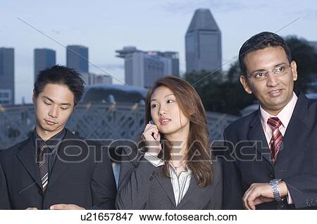 Stock Photo - Businesswoman standing with two businessmen and talking on a mobile phone, Singapore. Fotosearch