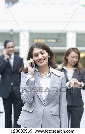 Stock Photo - Portrait of a businesswoman talking on a mobile phone with two business executives standing in the background. Fotosearch