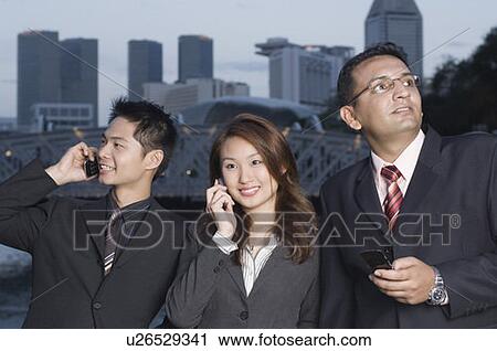 Stock Image - Two businessmen and a businesswoman standing together and using mobile phones, Singapore. Fotosearch