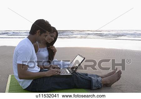Stock Image - Side profile of a young man sitting on a picnic blanket with a young woman and using a laptop. Fotosearch