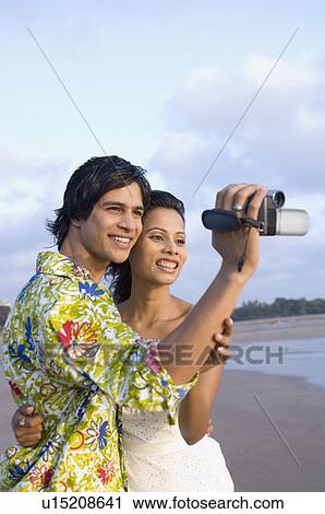 Stock Image - Young man holding a home video camera and a young woman embracing him. Fotosearch