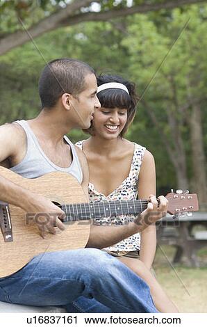 Stock Image - Young man sitting with his girlfriend and playing a guitar. Fotosearch