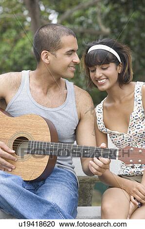 Young man sitting with his girlfriend and playing a guitar View Large Photo Image Stock Image - Young man sitting with his girlfriend and playing a guitar. Fotosearch