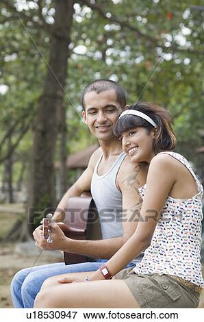 Young man sitting with his girlfriend and playing a guitar View Large Photo Image Stock Photo - Young man sitting with his girlfriend and playing a guitar. Fotosearch