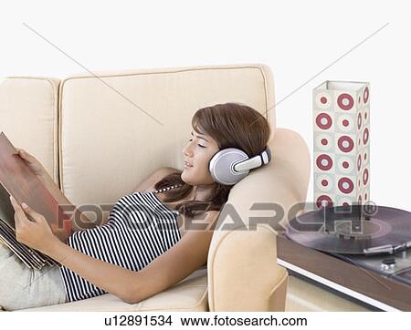 Picture - Side profile of a young woman reclining on a couch wearing headphones and listening to music. Fotosearch